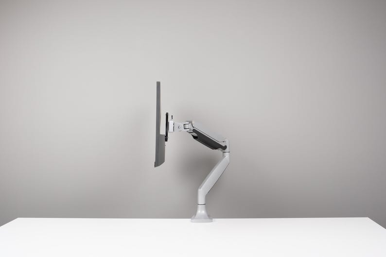 May include: A grey computer monitor mounted on a white and grey adjustable arm. The arm is attached to a white base, allowing for flexible positioning. The monitor is angled to the side.