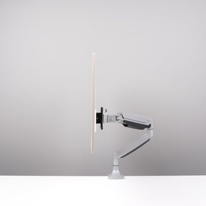 May include: A light gray monitor arm with a black mounting bracket supports a rectangular screen. The arm is attached to a white surface. The background is a light gray.
