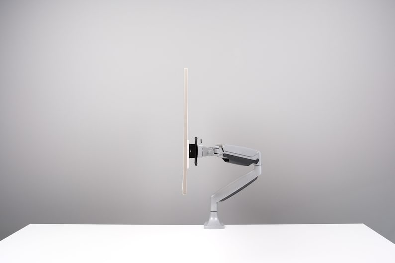 May include: A light gray monitor arm with a black mounting bracket supports a rectangular screen. The arm is attached to a white surface. The background is a light gray.