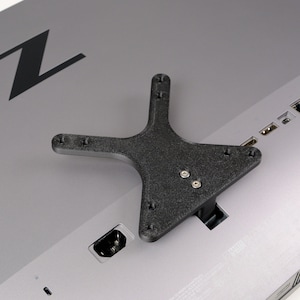 May include: A black X-shaped VESA mount adapter plate attached to the back of a silver computer monitor. The monitor features a large black Z logo on the upper left side and various input ports along the right edge, including USB and HDMI slots.