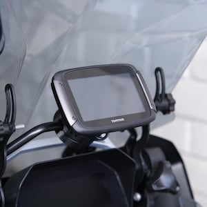 May include: A motorcycle GPS unit with a screen displaying the TomTom logo. The device is mounted on the handlebars, with a clear windshield in the background. The unit is black and grey.