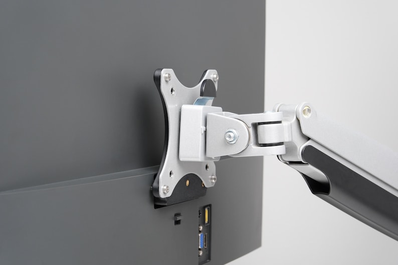 May include: A silver and black monitor arm is attached to the back of a dark grey computer monitor. The arm is designed to hold the monitor in place and allow for adjustments. The monitor has several ports.