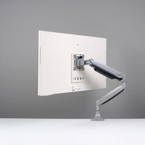 May include: A white computer monitor mounted on a gray adjustable arm. The monitor's back is visible, showing mounting hardware and a textured surface. The arm is attached to a base, allowing for flexible positioning.