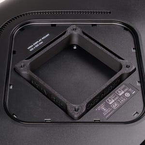 Puede incluir: Primer plano de un soporte de pantalla VESA negro, 100 x 100 mm, instalado en un monitor negro. El soporte tiene forma cuadrada con esquinas redondeadas y una abertura central. El monitor tiene una ventilación y una pequeña etiqueta.