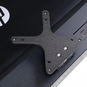 May include: A black metal bracket with multiple screw holes is attached to the back of a black computer monitor. The HP logo is visible in the upper left corner. The bracket has a unique shape with multiple arms.