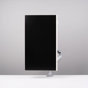 May include: A modern computer monitor with a black screen and a white frame. The monitor is mounted on a silver arm and base, and is positioned on a white desk against a gray background.