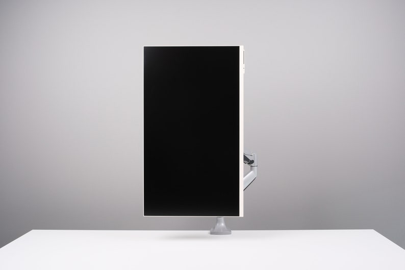 May include: A modern computer monitor with a black screen and a white frame. The monitor is mounted on a silver arm and base, and is positioned on a white desk against a gray background.
