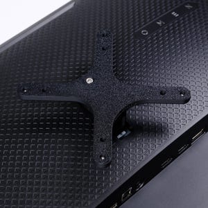 Pode incluir: Um suporte de montagem preto texturizado fixado na parte traseira de um monitor de computador preto. O monitor tem a palavra "OMEN" impressa. O suporte tem a forma de uma cruz.