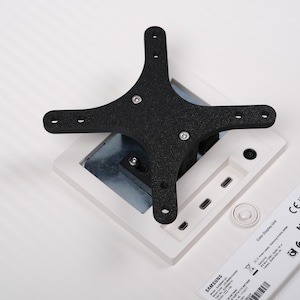 May include: A black, four-armed bracket with mounting holes is attached to a white monitor. The monitor has a white frame and a silver metal backing. A label with text and a QR code is visible.