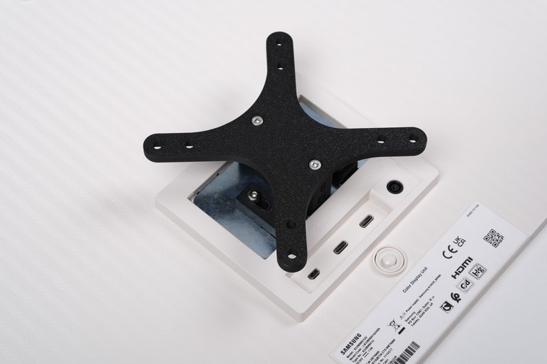 May include: A black, four-armed bracket with mounting holes is attached to a white monitor. The monitor has a white frame and a silver metal backing. A label with text and a QR code is visible.