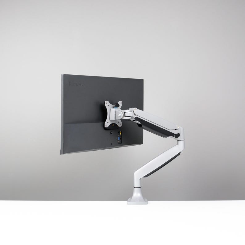 May include: A white and silver monitor arm is attached to the back of a dark grey computer monitor. The arm is designed to hold the monitor in place. The monitor and arm are on a white surface.