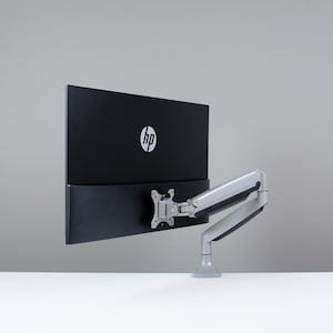 Puede incluir: Un monitor de ordenador HP negro con el logotipo de HP en blanco. El monitor está montado en un brazo ajustable plateado y gris. El brazo está fijado a una superficie blanca.