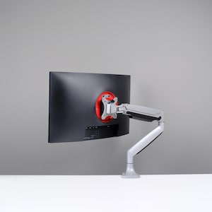 Puede incluir: Un monitor de ordenador negro montado en un brazo ajustable plateado y gris. La parte trasera del monitor es visible, con un soporte de montaje circular rojo. El brazo está fijado a una superficie blanca, con un fondo gris.