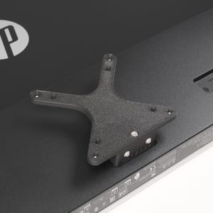 May include: A black, star-shaped bracket is mounted on the back of a black HP monitor. The bracket has multiple screw holes and a textured surface. The HP logo is visible on the monitor.