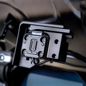 Può includere: Primo piano di un connettore nero con più pin, montato su una staffa in plastica nera. Il connettore è collegato a un filo nero.