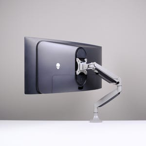 May include: A dark gray computer monitor with a white Alienware logo is mounted on a white and gray adjustable arm. The monitor is positioned against a light gray background, showcasing its design and mounting system.