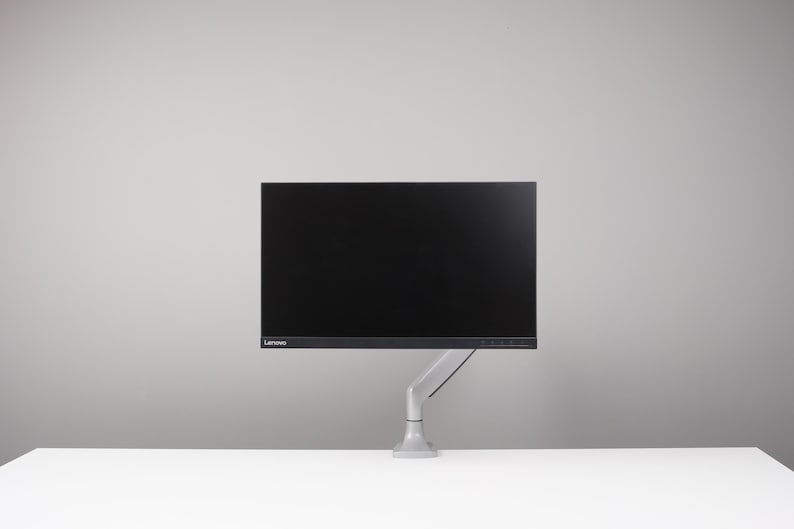 May include: A black Lenovo computer monitor mounted on a grey adjustable arm. The monitor is on a white surface against a grey background. The monitor is rectangular with a black screen.