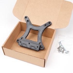 May include: A black plastic bracket with several holes, presented inside an open cardboard box. A small bag of hardware, including screws and an Allen key, is visible. The box is on a white surface.