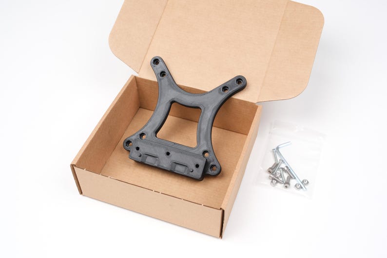 May include: A black plastic bracket with several holes, presented inside an open cardboard box. A small bag of hardware, including screws and an Allen key, is visible. The box is on a white surface.