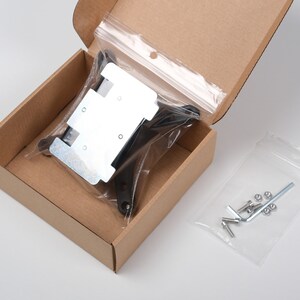 May include: A cardboard box containing hardware components. Inside, a clear plastic bag holds a metal bracket and black plastic pieces. Another bag contains screws, nuts, and an Allen wrench. The box is open, revealing the contents.