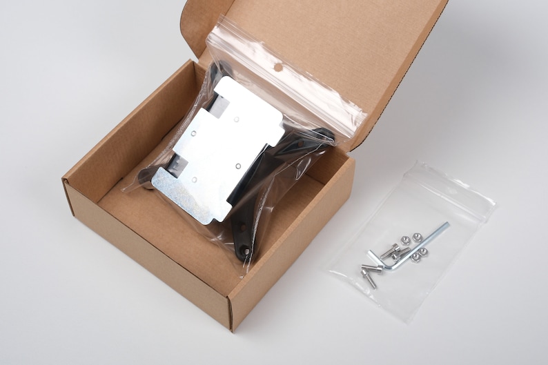 May include: A cardboard box containing hardware components. Inside, a clear plastic bag holds a metal bracket and black plastic pieces. Another bag contains screws, nuts, and an Allen wrench. The box is open, revealing the contents.