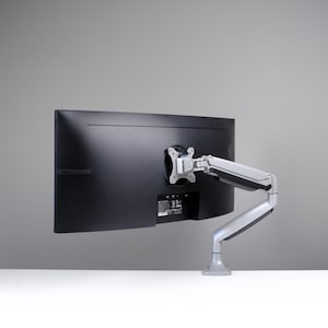 Puede incluir: Un monitor de ordenador negro montado en un brazo ajustable plateado. El monitor está colocado sobre un fondo gris, con el brazo extendiéndose desde la parte trasera de la pantalla. La base del brazo descansa sobre una superficie blanca.