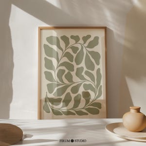 Op de afbeelding: Een ingelijste verticale wandprint met een saliegroen abstract botanisch bladpatroon op een lichtbeige achtergrond. De lijst is in een lichte houtkleur. De tekst FIRUM STUDIO met een klein bloemicoon staat onderaan in het midden.