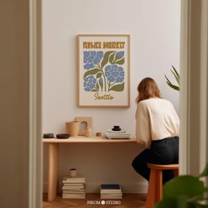May include: A framed print with a retro design featuring a stylized illustration of blue flowers and green leaves. The text "FLOWER MARKET Seattle" is printed in brown letters on a white background.