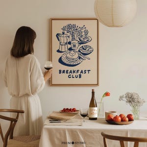 Könnte beinhalten: Ein gerahmter Wandkunstdruck mit einer dunkelblauen Strichzeichnung einer Frühstücksszene mit Mokkakanne, Pfannkuchen und einem Croissant auf cremefarbenem Hintergrund, mit dem Text BREAKFAST CLUB in dunkelblauen Großbuchstaben darunter.