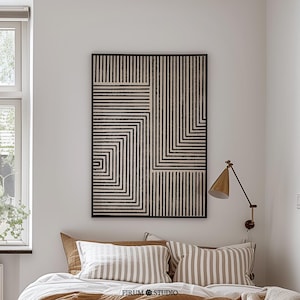 Può includere: Un'opera d'arte murale rettangolare caratterizzata da un motivo geometrico nero e beige di linee verticali e orizzontali che formano quadrati annidati. L'opera è incorniciata da un sottile bordo nero ed è esposta su una parete bianca sopra un letto.