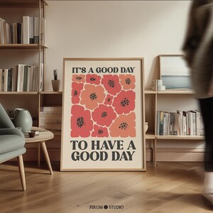 May include: A rectangular framed wall art print featuring a cream background with a central cluster of stylized red and coral flowers with dark centers. The text IT'S A GOOD DAY TO HAVE A GOOD DAY is printed in bold black capital letters.