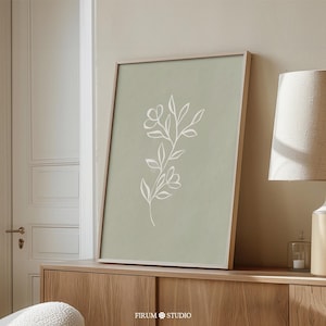 May include: A framed wall art print featuring a sage green background with a white line drawing of a floral stem with leaves and two blossoms. The artwork is displayed in a light-colored wooden frame, positioned on a wooden surface.