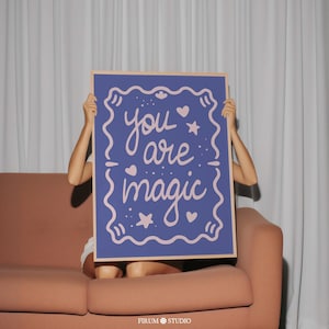 May include: A rectangular wall art print featuring a deep blue background with a light pink decorative border. The text "you are magic" is written in a cursive, light pink font in the center, surrounded by small light pink stars and hearts.