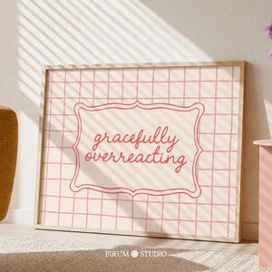 Pode incluir: Uma impressão de arte de parede retangular emoldurada com um fundo creme claro e um padrão de grade rosa. Centralizado está uma moldura decorativa rosa recortada contendo o texto vermelho em cursivo "gracefully overreacting" em fonte legível.
