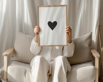 May include: A rectangular wooden frame holding a white print with a centered black heart graphic. The frame is held up, obscuring the face of the person behind it. The print features a simple, solid black heart shape on a plain white background.