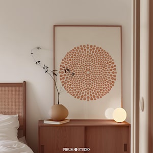 Puede incluir: Una lámina de arte enmarcada con un gran patrón circular tipo mandala compuesto por pequeños pétalos de color terracota sobre un fondo crema. La lámina se exhibe sobre un mueble de madera con un jarrón y una pequeña lámpara de globo brillante.