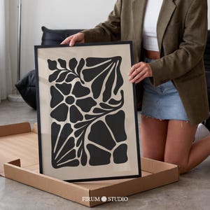May include: Black framed art print featuring a floral design in black against a beige background. The artwork is held in a cardboard box. The text "FIRUM STUDIO" is printed at the bottom of the box.