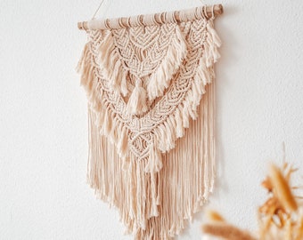 May include: A beige macrame wall hanging with a geometric pattern and fringe. The hanging is made of natural fibers and has a bohemian style.