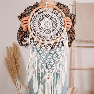 Puede incluir: Un atrapasue&ntilde;os de crochet blanco con cuentas de madera y plumas blancas que cuelgan de un aro de madera.