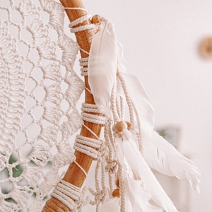 Puede incluir: Un atrapasue&ntilde;os de macram&eacute; blanco con plumas blancas y cuentas de madera. El atrapasue&ntilde;os est&aacute; colgado de un aro de madera.