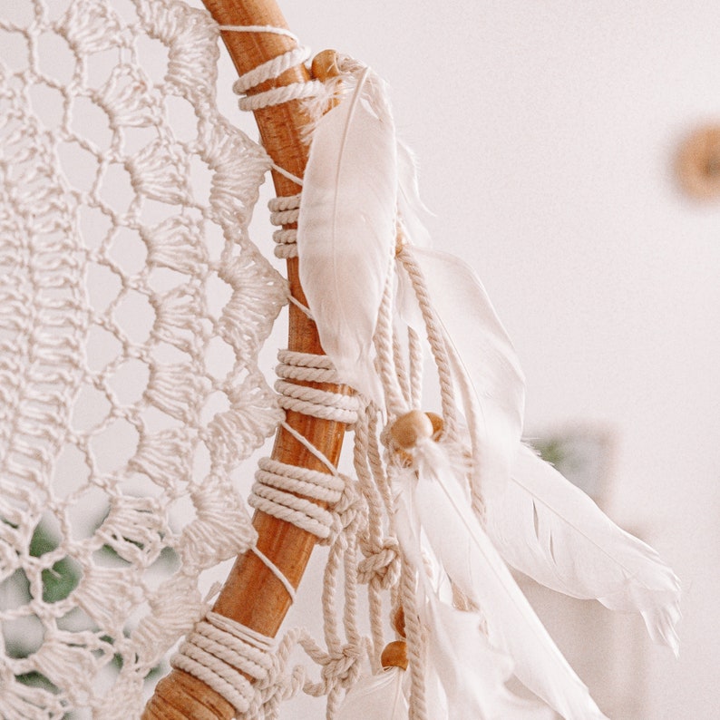 Puede incluir: Un atrapasue&ntilde;os de macram&eacute; blanco con plumas blancas y cuentas de madera. El atrapasue&ntilde;os est&aacute; colgado de un aro de madera.