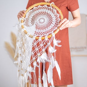 Puede incluir: Un atrapasue&ntilde;os blanco con un aro de madera, plumas blancas y macram&eacute; blanco. El atrapasue&ntilde;os est&aacute; colgado de una varilla de madera.