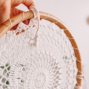Puede incluir: Un atrapasue&ntilde;os de crochet blanco con un aro de madera y un cord&oacute;n blanco que cuelga de la parte superior.