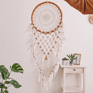 Puede incluir: Un atrapasue&ntilde;os blanco y marr&oacute;n con un aro de madera y plumas blancas que cuelgan de &eacute;l. El atrapasue&ntilde;os est&aacute; colgado de una pared blanca.