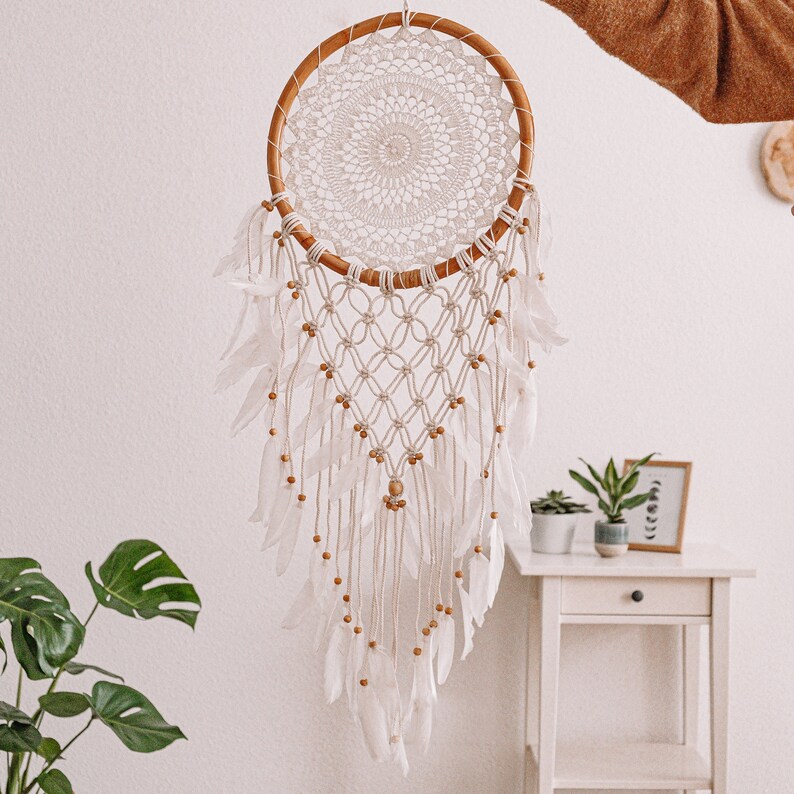 Puede incluir: Un atrapasue&ntilde;os blanco y marr&oacute;n con un aro de madera y plumas blancas que cuelgan de &eacute;l. El atrapasue&ntilde;os est&aacute; colgado de una pared blanca.