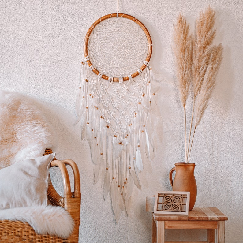 Puede incluir: Un atrapasue&ntilde;os de macram&eacute; blanco con un aro de madera y plumas blancas que cuelgan de &eacute;l. El atrapasue&ntilde;os est&aacute; colgado en una pared blanca con un jarr&oacute;n de hierba de la pampa seca a la derecha.