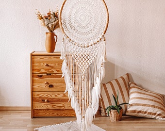 May include: A large, handmade dreamcatcher with a circular wooden frame and intricate white macrame detailing. White feathers and wooden beads hang from the bottom. The dreamcatcher is suspended in front of a wooden dresser and decorative pillows.