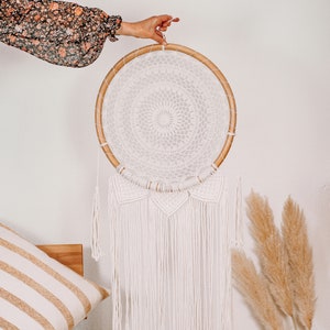 Puede incluir: Un atrapasueños de macramé blanco con un aro de madera y flecos blancos que cuelgan de la parte inferior. El atrapasueños está colgado de una pared blanca.