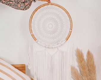 May include: A white macrame dream catcher with a wooden hoop and white fringe hanging from the bottom. The dream catcher is hanging from a white wall.
