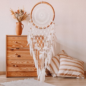 Puede incluir: Un atrapasue&ntilde;os de macram&eacute; blanco con plumas que cuelgan de un aro de madera. El atrapasue&ntilde;os est&aacute; colgado de una pared blanca sobre una c&oacute;moda de madera con tres cajones.